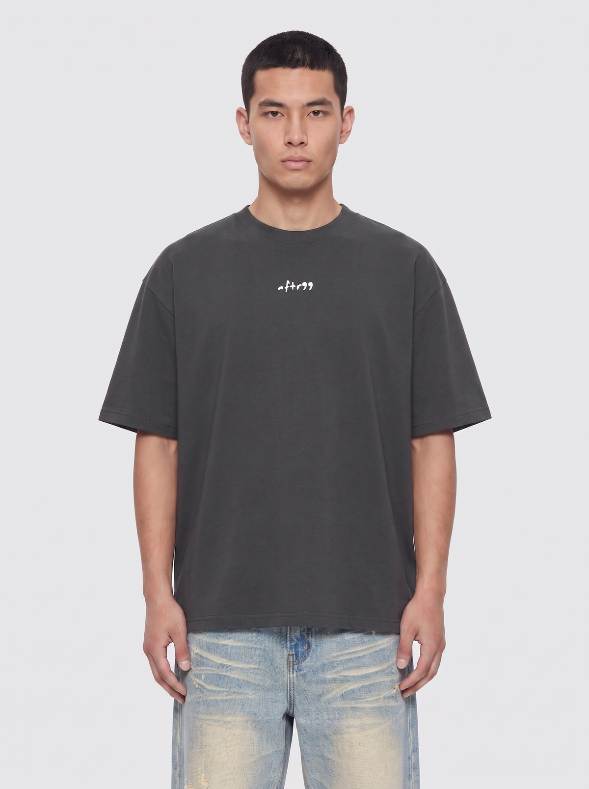 A man wearing a dark gray oversized t-shirt with a small white graphic on the chest, paired with light-wash distressed jeans, against a light gray background.