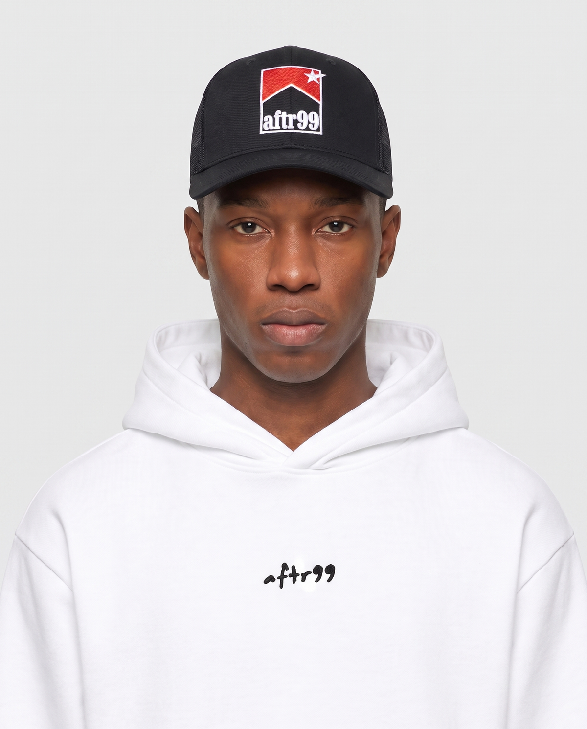 aftr99 | A model wearing a black baseball cap with a red, white, and black 'afr99' logo, paired with a white hoodie featuring a small black 'afr99' logo on the chest, against a light gray background.