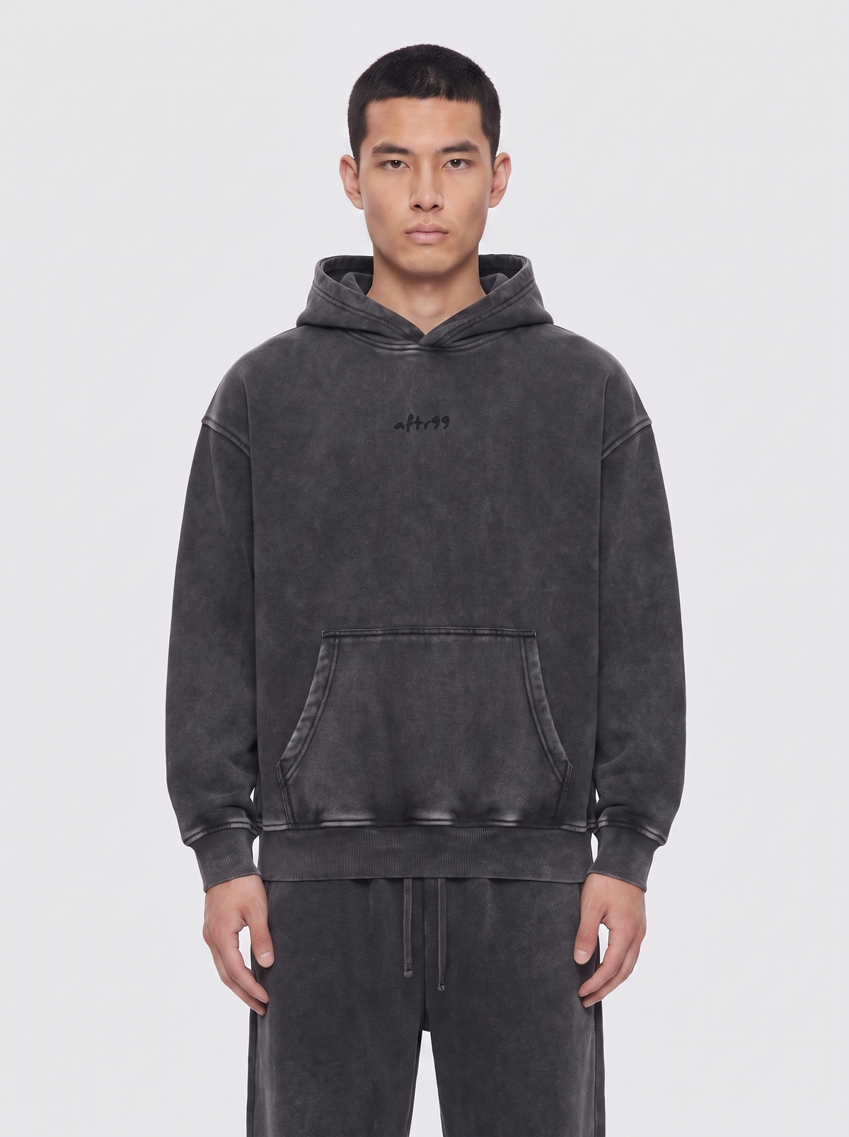 aftr99 | A man wearing a dark gray washed hoodie with a small black embroidered logo on the chest, paired with matching dark gray sweatpants, against a light gray studio background.
