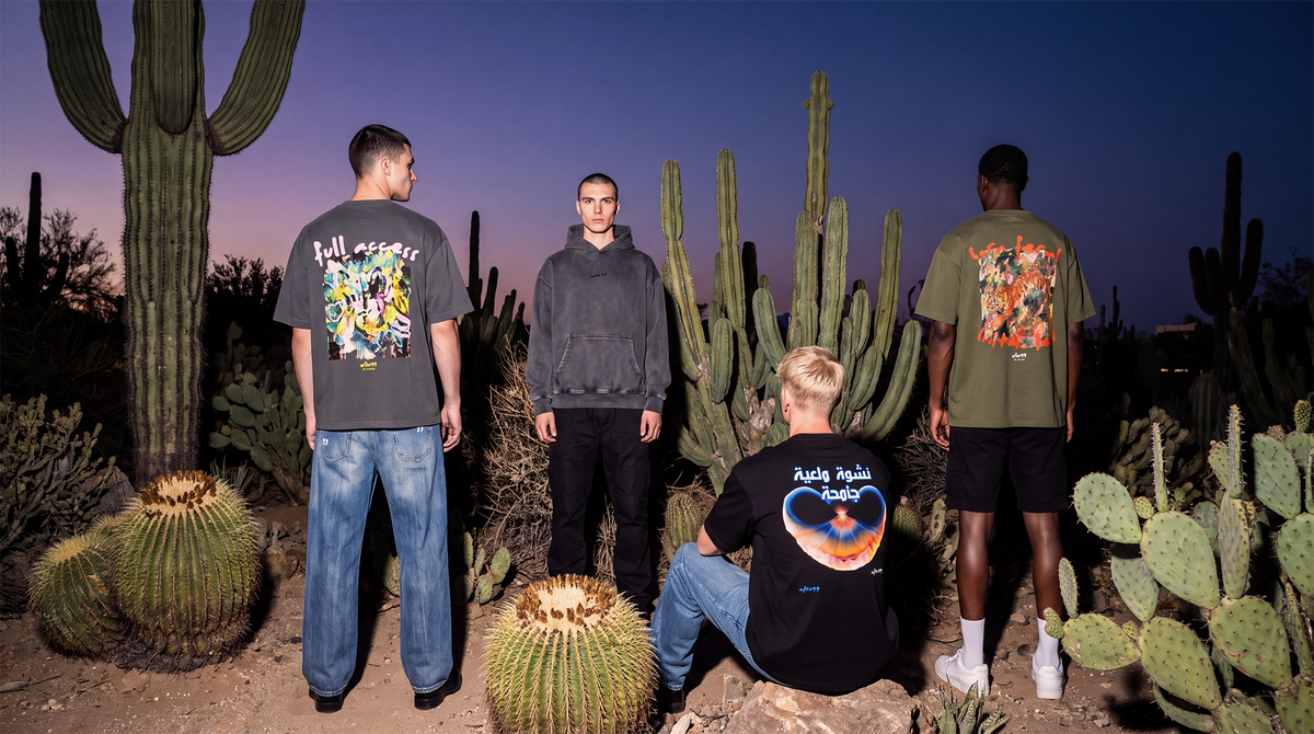 aftr99 collective | Four men standing outdoors in a desert at dusk wearing matching graphic t-shirts with large back prints. The shirts feature colorful designs including a yellow and blue graphic on one, a dark graphic on another, and a green shirt with a red and orange graphic on the third.