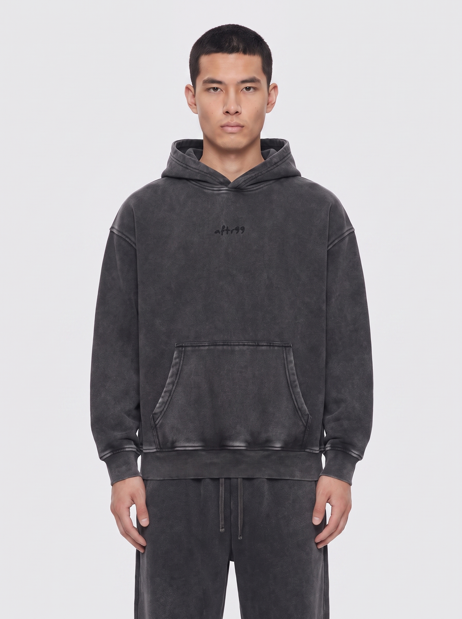 aftr99 | A man wearing a dark gray washed hoodie with a small black embroidered logo on the chest, paired with matching dark gray sweatpants, against a light gray studio background.