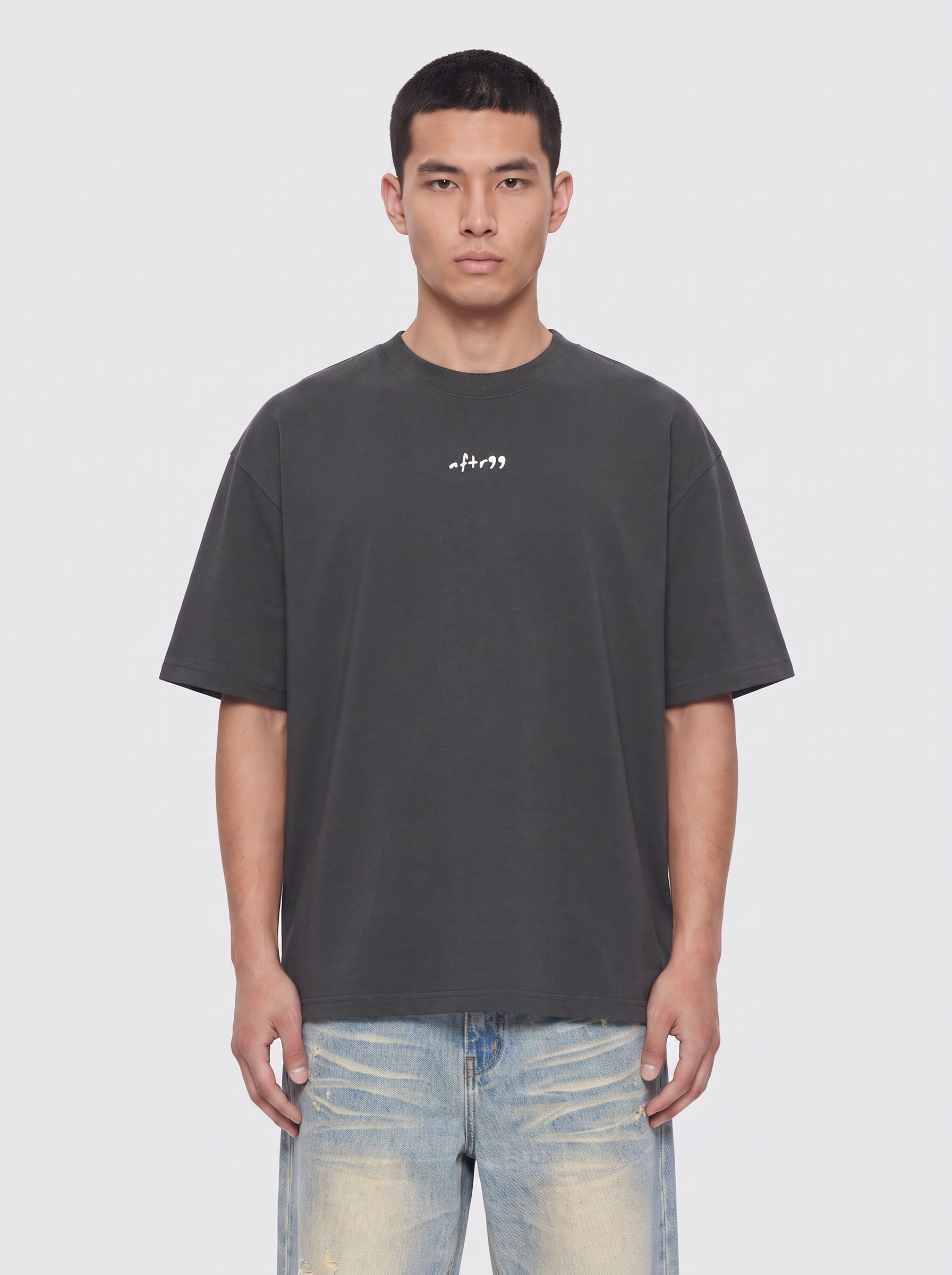 A man wearing a dark gray oversized t-shirt with a small white graphic on the chest, paired with light-wash distressed jeans, against a light gray background.