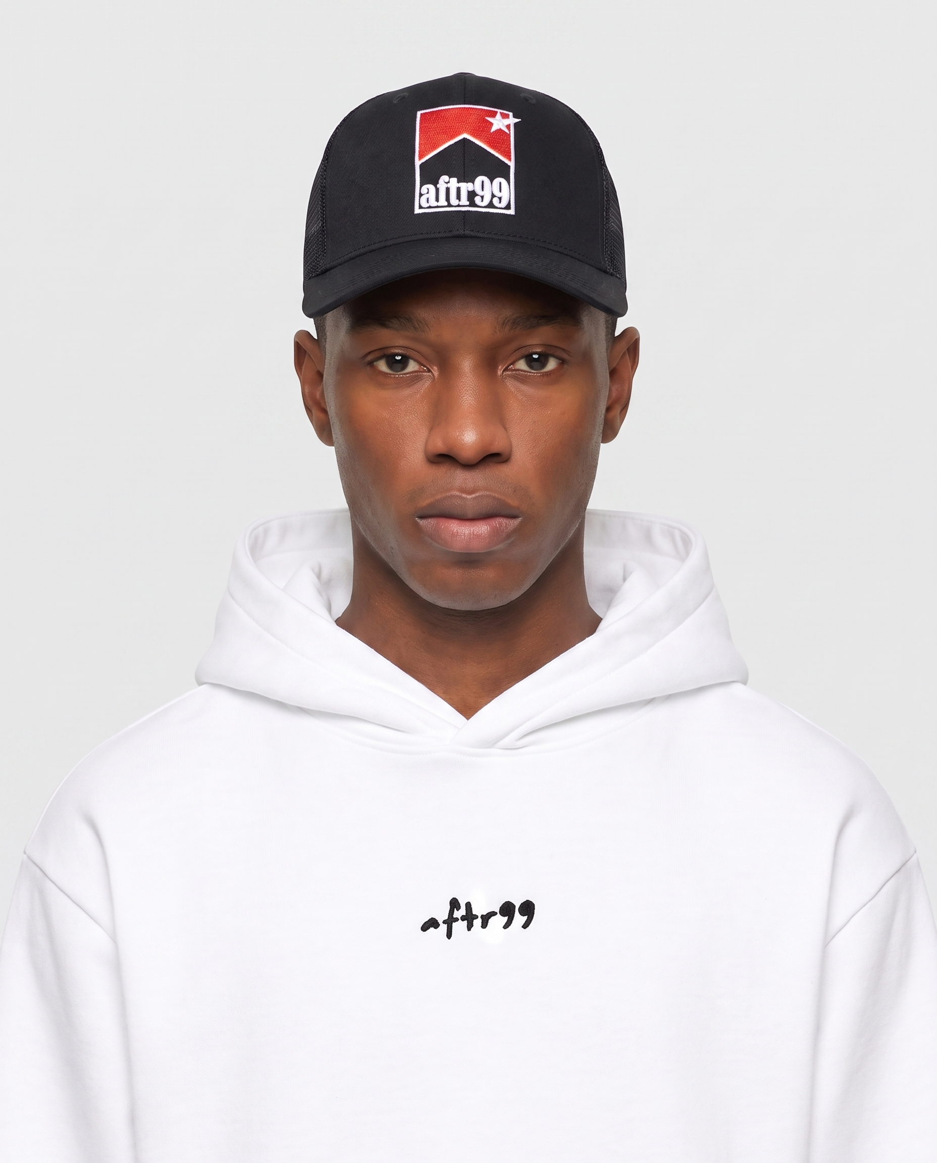 aftr99 | A model wearing a black baseball cap with a red, white, and black 'afr99' logo, paired with a white hoodie featuring a small black 'afr99' logo on the chest, against a light gray background.