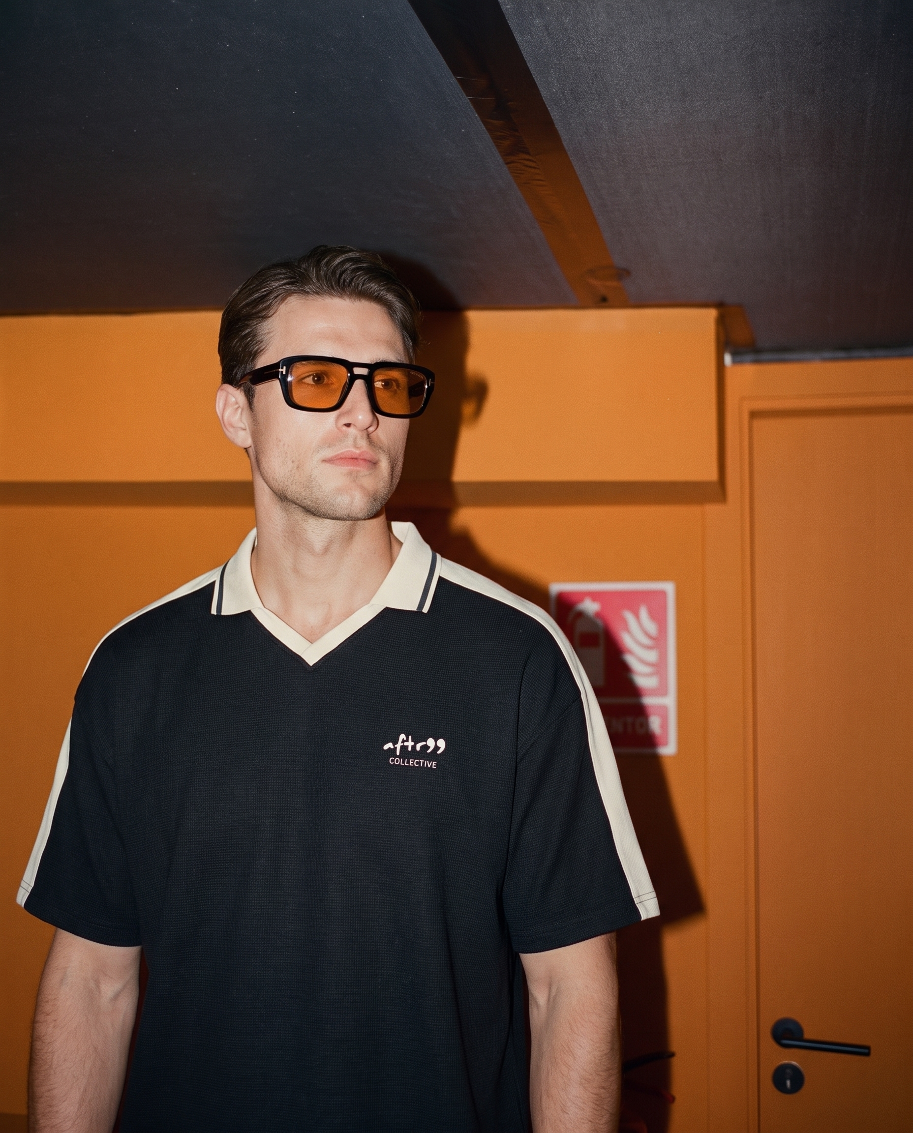 aftr99 collective | A man wearing black-framed tinted glasses and a black short-sleeve polo shirt with cream shoulder stripes and a small white logo on the chest, standing in front of an orange wall.
